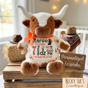 May include: A plush longhorn cow toy with white and brown fur, featuring an orange ribbon around its neck. The chest is embroidered with the name Marco William, birth details including 7 pounds 1 ounce, 20 inches, 3:06 PM, and October 14, 2025.