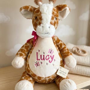 May include: A plush giraffe stuffed animal with brown and white patterned fur. The giraffe has a white belly embroidered with the name "Lucy" in pink, along with pink butterfly designs. A pink ribbon is tied around its neck. A tag reads "Handmade with Love."