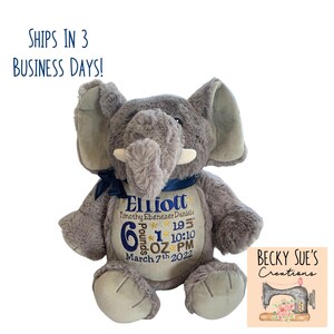 May include: A gray plush elephant toy with a dark blue ribbon around its neck. The elephant's belly features embroidered text including the name Elliott, Timothy Ebenezer Daniels, birth date March 7th 2022, and birth stats 6 pounds 1 ounce.