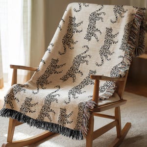 May include: A beige throw blanket featuring a black leopard print, draped over a wooden rocking chair. The blanket has a fringed edge in black and multicoloured threads. The leopard pattern is repeated across the blanket.