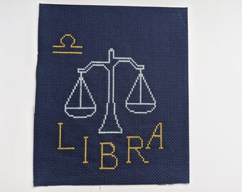 May include: A navy blue cross-stitch embroidery with a white outline of a scale and the word "LIBRA" in gold thread.
