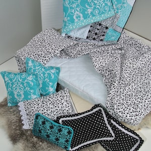 May include: A doll-sized bed with a blue and white patterned quilt, a black and white patterned sheet, and several pillows with different patterns. The bed is on a white surface with a fluffy gray rug.