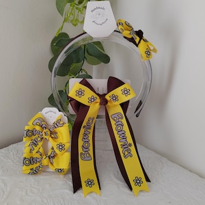 Può includere: Un cerchietto giallo e marrone scuro con un grande fiocco che riporta la scritta Brownies in viola chiaro, decorato con motivi a margherite bianche. Un fiocco più piccolo abbinato è fissato al cerchietto e un fiocco per capelli separato è incluso.