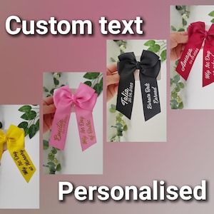 May include: Four hair bows in yellow, pink, black, and dark pink, each featuring custom white or gold text. The ribbons display names, dates, and phrases like My 1st Day at Nursery, My 1st Day of Ballet, Karate Belt Earned, and My 1st Day at school.