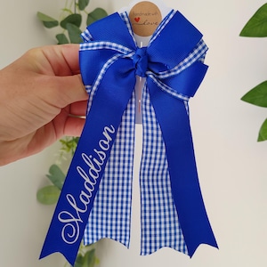 May include: A royal blue hair bow featuring a white and blue gingham pattern on the ribbon tails. The name Maddison is printed in white script on one of the long ribbon tails. The bow is attached to a white card with a handmade with love tag.
