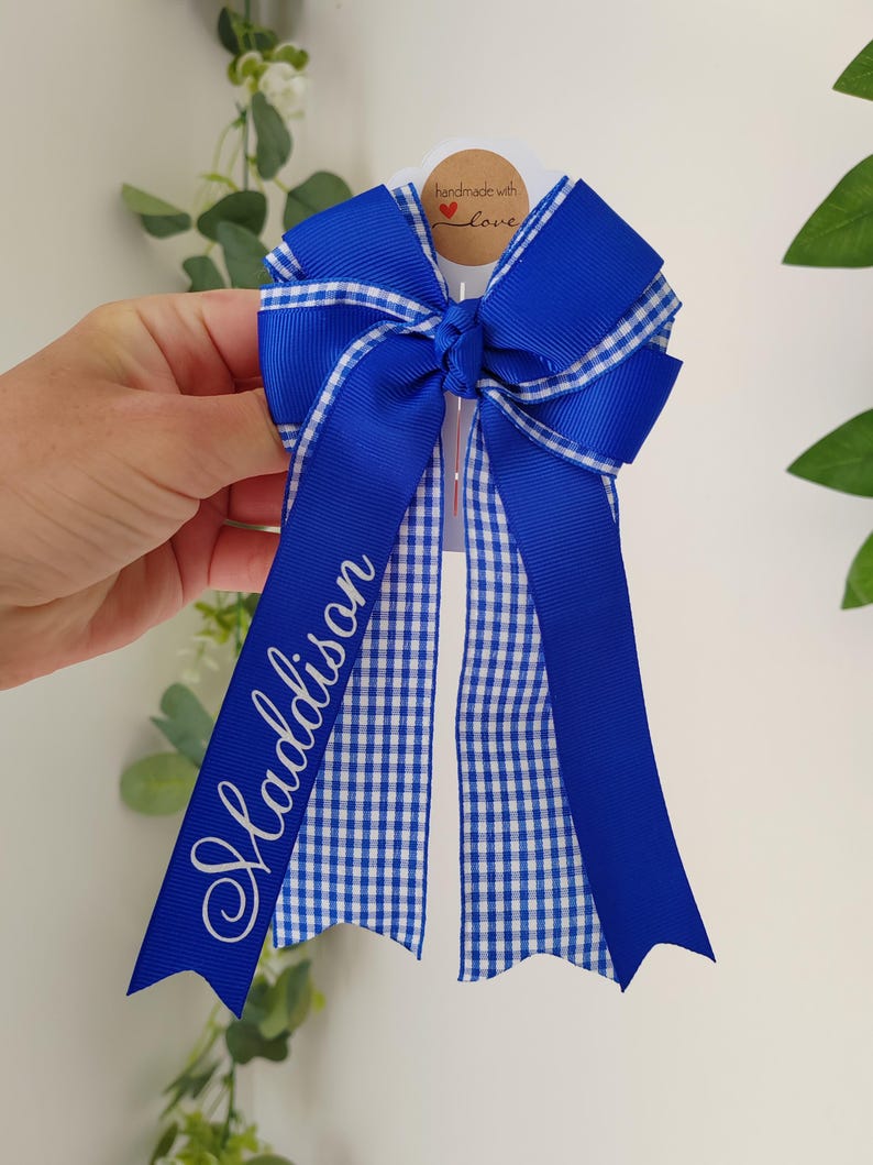 May include: A royal blue hair bow featuring a white and blue gingham pattern on the ribbon tails. The name Maddison is printed in white script on one of the long ribbon tails. The bow is attached to a white card with a handmade with love tag.