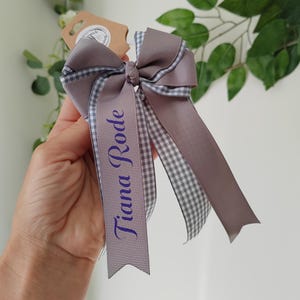 May include: A hair bow made of solid mauve grosgrain ribbon layered with a grey and white gingham patterned ribbon. The bow features two long tails, one of which is printed with the name Tiana Rode in a dark purple script font.