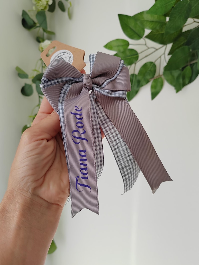May include: A hair bow made of solid mauve grosgrain ribbon layered with a grey and white gingham patterned ribbon. The bow features two long tails, one of which is printed with the name Tiana Rode in a dark purple script font.
