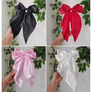May include: Four satin hair bows in black, red, pink, and white. Each bow features a large knot and long, flowing tails. The bows are displayed against a backdrop of green leaves.