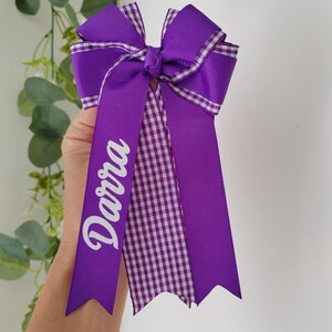 May include: A purple hair bow featuring a layered design with solid purple ribbon and a purple and white gingham checkered pattern. The bow has long, pointed tails, and the name Darra is printed in white script lettering on one of the ribbons.