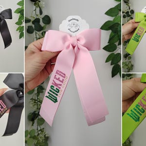 Könnte beinhalten: Eine Auswahl handgefertigter Haarschleifen in verschiedenen Farben, darunter Schwarz, Rosa und Grün. Jede Schleife zeigt die Wörter "WICKED" und "FOR GOOD" in Glitzerbuchstaben. Die Schleifen sind an einem weißen Etikett mit einem Logo befestigt.