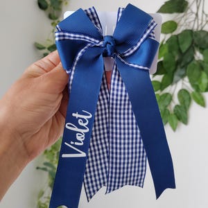 May include: A blue hair bow featuring a combination of solid navy blue ribbon and a blue and white gingham checkered pattern. The bow has long, flowing tails, and the name Violet is printed in white script lettering on one of the long ribbons.
