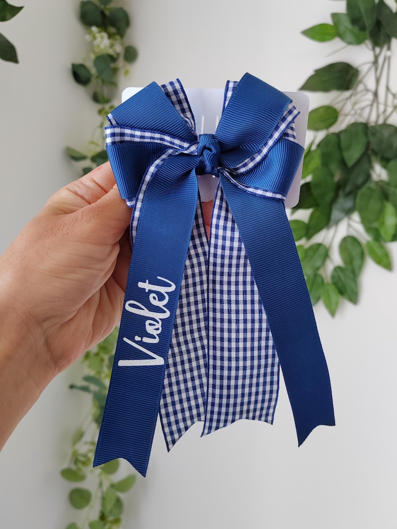 May include: A blue hair bow featuring a combination of solid navy blue ribbon and a blue and white gingham checkered pattern. The bow has long, flowing tails, and the name Violet is printed in white script lettering on one of the long ribbons.