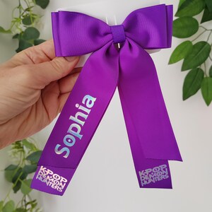 May include: A vibrant purple hair bow with long streamers. The word "Sophia" is printed in a light blue, iridescent font on one streamer. The other streamer has the text "KPOP DEMON HUNTERS" in a white font.