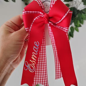May include: A red hair bow made of grosgrain ribbon with a white and red gingham check trim. The bow features two long tails, one of which is personalized with the name Esmae in a cursive, iridescent script. A small tag reads Handmade.