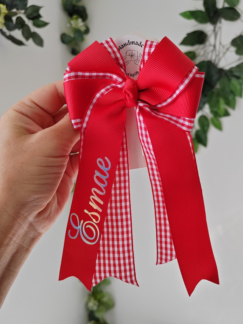 May include: A red hair bow made of grosgrain ribbon with a white and red gingham check trim. The bow features two long tails, one of which is personalized with the name Esmae in a cursive, iridescent script. A small tag reads Handmade.