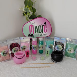May include: A selection of witch-themed items, including pink and black cauldrons, small bottles, and bags of colourful ingredients. The sign says "The Magic is alive". The items are arranged on a white surface.
