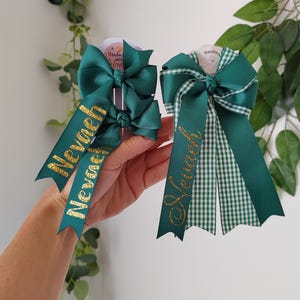 May include: Two dark green hair bows made of grosgrain ribbon. The left bow features two smaller bows with the name Nevaeh in gold glitter. The right bow is a larger layered design with a green and white gingham pattern and the name Nevaeh in gold.