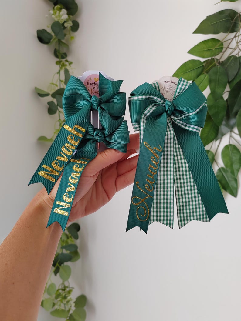 May include: Two dark green hair bows made of grosgrain ribbon. The left bow features two smaller bows with the name Nevaeh in gold glitter. The right bow is a larger layered design with a green and white gingham pattern and the name Nevaeh in gold.