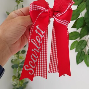 May include: A red hair bow made of grosgrain ribbon with a white and red gingham pattern trim. The bow features the name Scarlett written in silver glitter script on one of the long, dangling ribbon tails, which have notched ends.