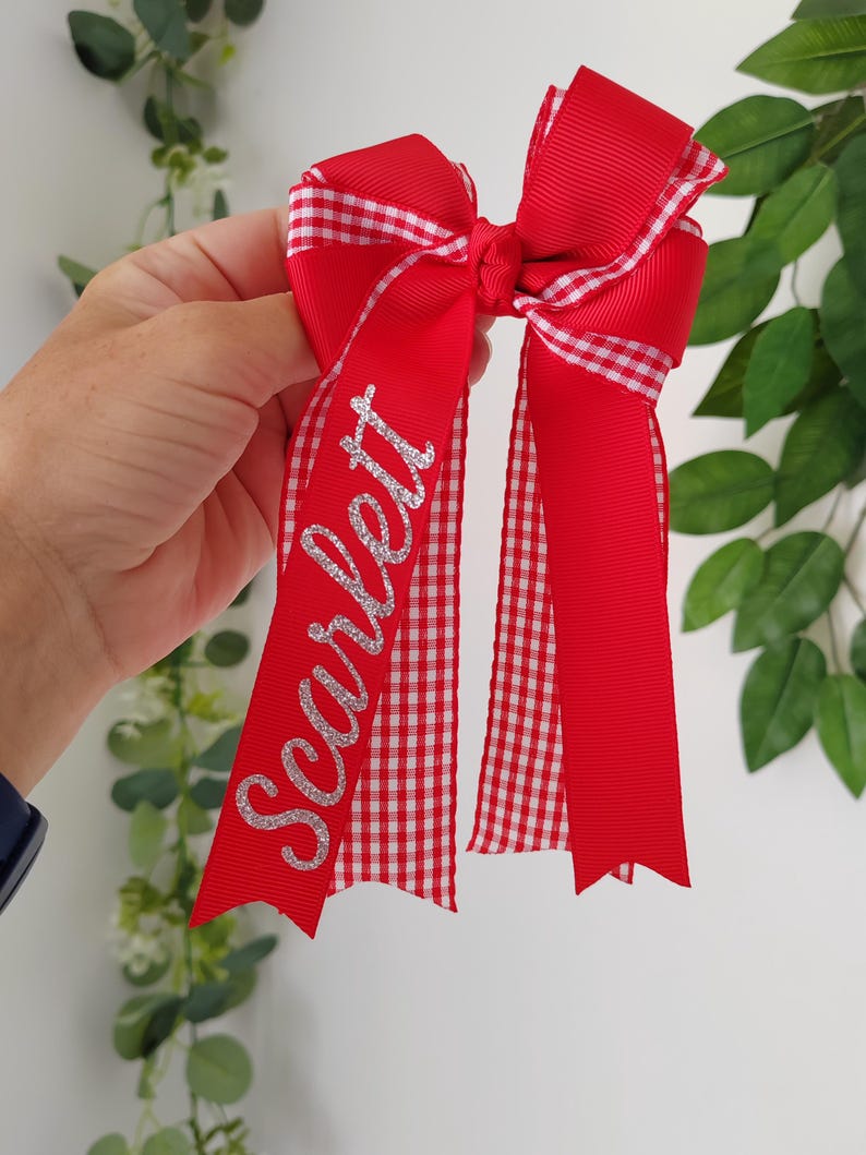 May include: A red hair bow made of grosgrain ribbon with a white and red gingham pattern trim. The bow features the name Scarlett written in silver glitter script on one of the long, dangling ribbon tails, which have notched ends.
