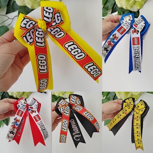 Pode incluir: Uma coleção de cinco laços de cabelo com vários temas LEGO e Ninjago. Os laços são feitos de fita de gorgorão nas cores amarelo, vermelho, azul e preto, com logotipos impressos, gráficos de personagens e a palavra Name visível.