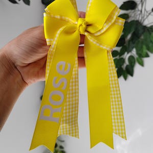 May include: A bright yellow hair bow constructed from solid yellow grosgrain ribbon layered with a white and yellow gingham patterned ribbon. The bow features two long tails, with the name Rose printed in white capital letters on the left tail.