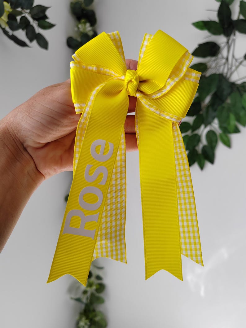 May include: A bright yellow hair bow constructed from solid yellow grosgrain ribbon layered with a white and yellow gingham patterned ribbon. The bow features two long tails, with the name Rose printed in white capital letters on the left tail.