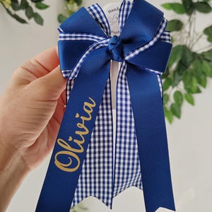 May include: A hair bow featuring layered navy blue and white gingham ribbon with solid navy blue ribbon accents. The bow is tied in a central knot and has two long, pointed tails. The name Olivia is printed in gold script on one of the tails.