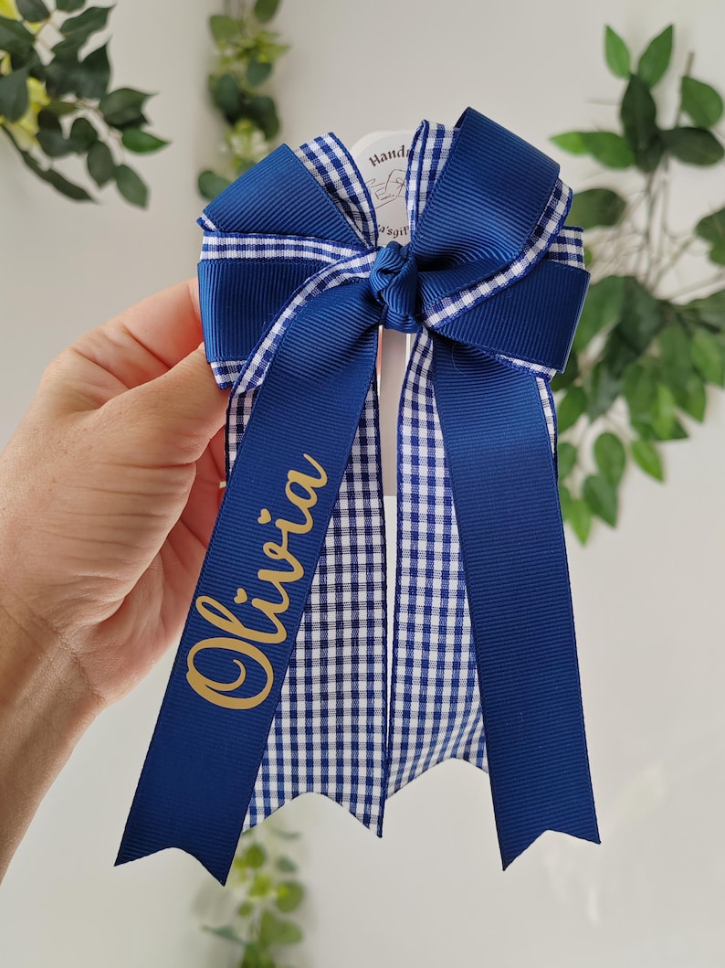 May include: A hair bow featuring layered navy blue and white gingham ribbon with solid navy blue ribbon accents. The bow is tied in a central knot and has two long, pointed tails. The name Olivia is printed in gold script on one of the tails.