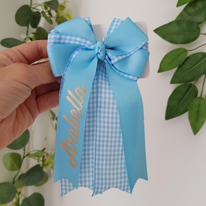 May include: A light blue hair bow made of grosgrain ribbon with a white and light blue gingham pattern. The bow features a central knot and long, dangling ribbon tails. The name Arabella is printed in gold script on one of the ribbon tails.
