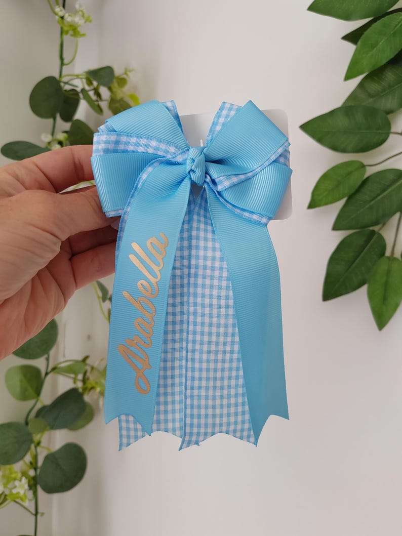 May include: A light blue hair bow made of grosgrain ribbon with a white and light blue gingham pattern. The bow features a central knot and long, dangling ribbon tails. The name Arabella is printed in gold script on one of the ribbon tails.