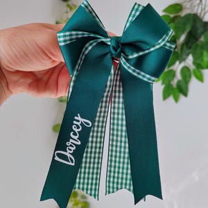 May include: A dark green grosgrain ribbon hair bow featuring a white and green gingham check pattern on the edges and center tails. The left tail is personalized with the name Darcey printed in white script lettering on the solid green ribbon.
