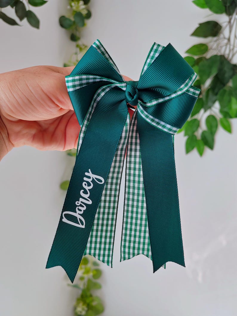May include: A dark green grosgrain ribbon hair bow featuring a white and green gingham check pattern on the edges and center tails. The left tail is personalized with the name Darcey printed in white script lettering on the solid green ribbon.