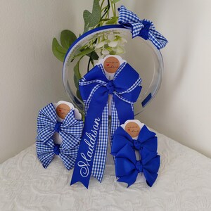 May include: A collection of royal blue hair accessories including a headband with a gingham bow, a large personalized bow with the name Maddison in white script, and two sets of smaller hair bows in solid royal blue and blue gingham patterns.