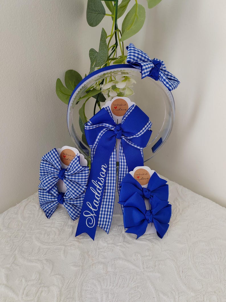 May include: A collection of royal blue hair accessories including a headband with a gingham bow, a large personalized bow with the name Maddison in white script, and two sets of smaller hair bows in solid royal blue and blue gingham patterns.
