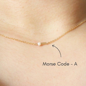 May include: A delicate gold necklace featuring a small, white bead and a horizontal bar. The necklace is worn around the neck and has a fine chain. The text "Morse Code - A" is next to the necklace.
