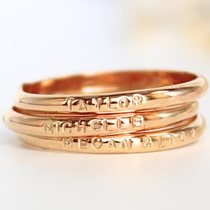 May include: A stack of four gold-colored rings. Each ring is engraved with a name: "TAYLOR", "MICHELLE", "MEGAN & LYNN". The rings are stacked together, creating a layered effect. The background is blurred, focusing attention on the rings.