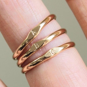 Puede incluir: Tres anillos finos de tono dorado apilados, cada uno con una pequeña sección central plana y pulida. El anillo superior está grabado con la letra A, el anillo central con la letra B y el anillo inferior con la letra C en fuente simple.