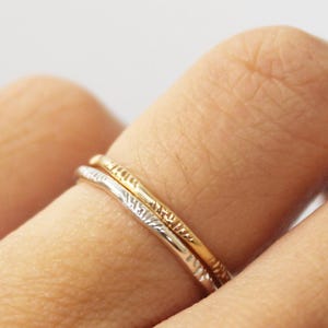 May include: A set of two thin stacking rings, one in a polished gold tone and one in a polished silver tone. Both rings feature a delicate, uneven, and textured surface along the entire circumference of the thin, rounded metal bands.