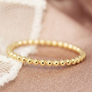 Puede incluir: Un delicado anillo de oro con una serie de pequeñas cuentas espaciadas uniformemente. El anillo se presenta sobre un fondo suave, de color beige rosado, con un toque de encaje visible a la izquierda. El diseño del anillo es sencillo y elegante.