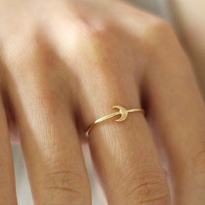 May include: A delicate gold-colored ring featuring a crescent moon design. The ring is thin and minimalist, with the moon shape as the central focus. The ring is worn on a finger, showcasing its simple elegance.