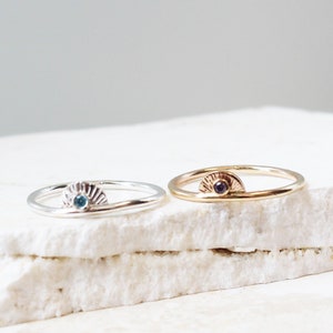 Puede incluir: Dos anillos delicados con un diseño de sol y una pequeña piedra preciosa en el centro. El anillo de la izquierda tiene una piedra preciosa azul, mientras que el anillo de la derecha tiene una piedra preciosa morada.