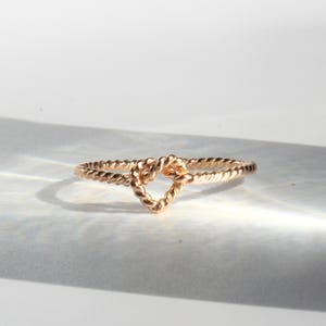 Op de afbeelding: Een roségouden ring met een gedraaide, touwachtige band die een centraal hartvormig element vormt. Het metaal heeft een gepolijste afwerking die het licht reflecteert en de ingewikkelde textuur van het gedraaide draadontwerp over de ring benadrukt.