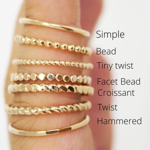 May include: A collection of seven gold-toned rings stacked on a finger, labelled as Simple, Bead, Tiny twist, Facet Bead, Croissant, Twist, and Hammered. Each ring features a distinct texture or shape, ranging from smooth bands to beaded and twisted designs.