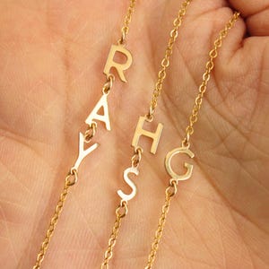 Peut inclure: Colliers dorés avec des pendentifs individuels en forme de lettres. Les lettres épellent des noms. Les colliers sont présentés sur un fond de couleur chair. Chaque collier a une chaîne fine.