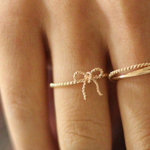 May include: Two gold rings on a finger. The ring on the index finger is a thin band. The ring on the middle finger is a thin band with a small bow design.