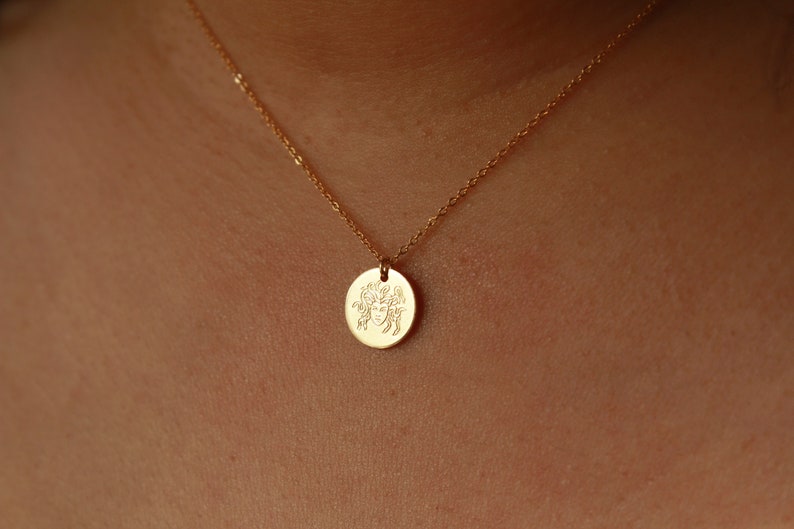 K&ouml;nnte beinhalten: Eine goldene Kettenkette mit einem runden Anh&auml;nger, der ein eingraviertes Bild eines Frauenkopfes mit einer Bl&auml;tterkrone zeigt.
