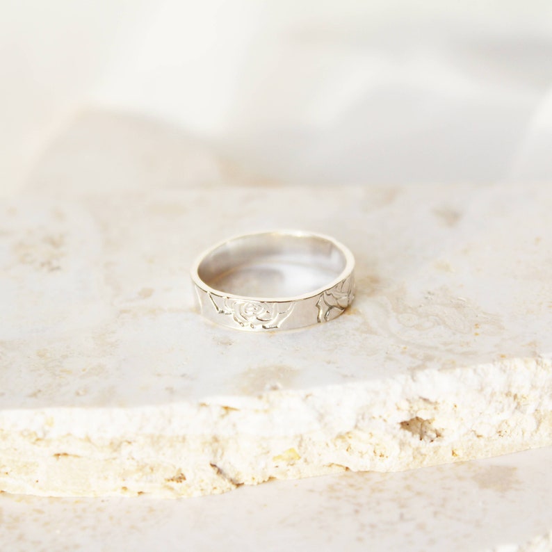 Puede incluir: Un anillo de plata con un dise&ntilde;o floral. El anillo presenta un patr&oacute;n grabado de rosas y hojas alrededor de la banda. Se muestra sobre una superficie clara y texturizada.