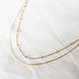 May include: Two gold necklaces, one with a delicate chain and the other with a chain featuring small beads.